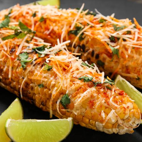 The image shows two pieces of Mexican street corn (elote) garnished with cheese, cilantro, and seasoning, with lime wedges on the side.