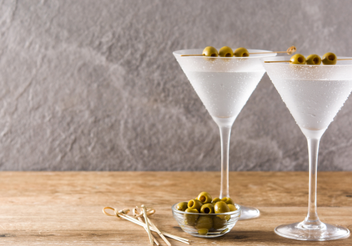 The image shows two martini glasses filled with a drink, each garnished with green olives. A small bowl of olives and some cocktail sticks are beside them.