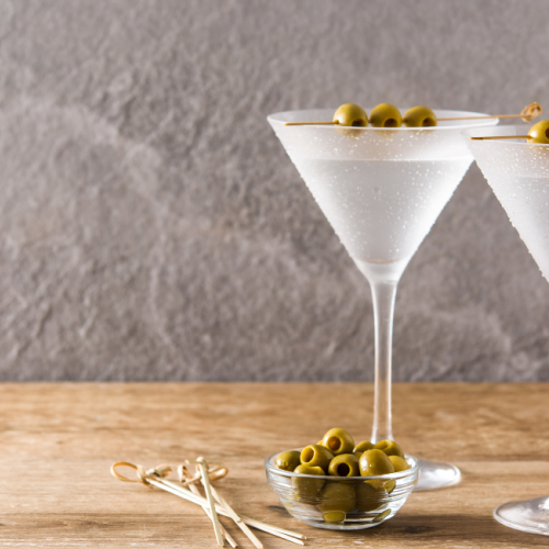 The image shows two martini glasses filled with a drink, each garnished with green olives. A small bowl of olives and some cocktail sticks are beside them.