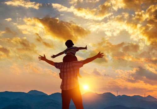 A person carrying a child on their shoulders with both having outstretched arms, silhouetted against a dramatic sunset sky and mountain landscape.