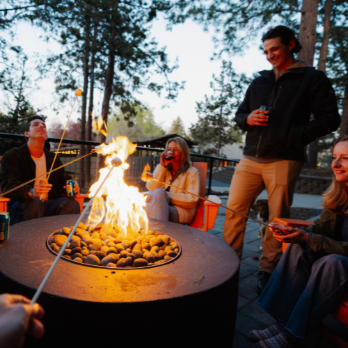 People are gathered around a fire pit, roasting marshmallows and enjoying drinks in an outdoor setting with trees.