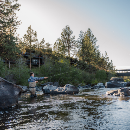 Guest fly fishing in the Deschutes River near Riverhouse Lodge, surrounded by lush trees, rocky banks, and blue skies—an iconic Bend adventure.