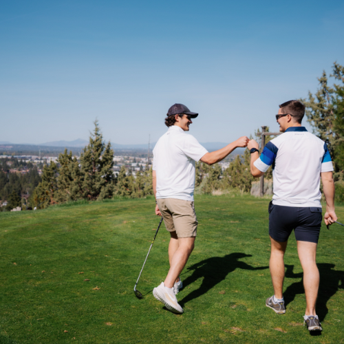 Two people on a golf course, giving each other a fist bump. One is holding a golf club. Trees and distant hills are in the background.