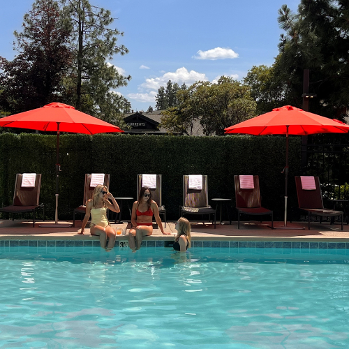 Guests lounging poolside under red umbrellas at Riverhouse Lodge, enjoying a sunny summer day with views of the river and pines.