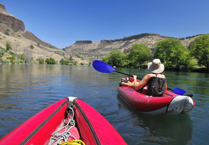 Two people kayaking on a serene river surrounded by hills and greenery under a clear blue sky, with one person in the foreground.