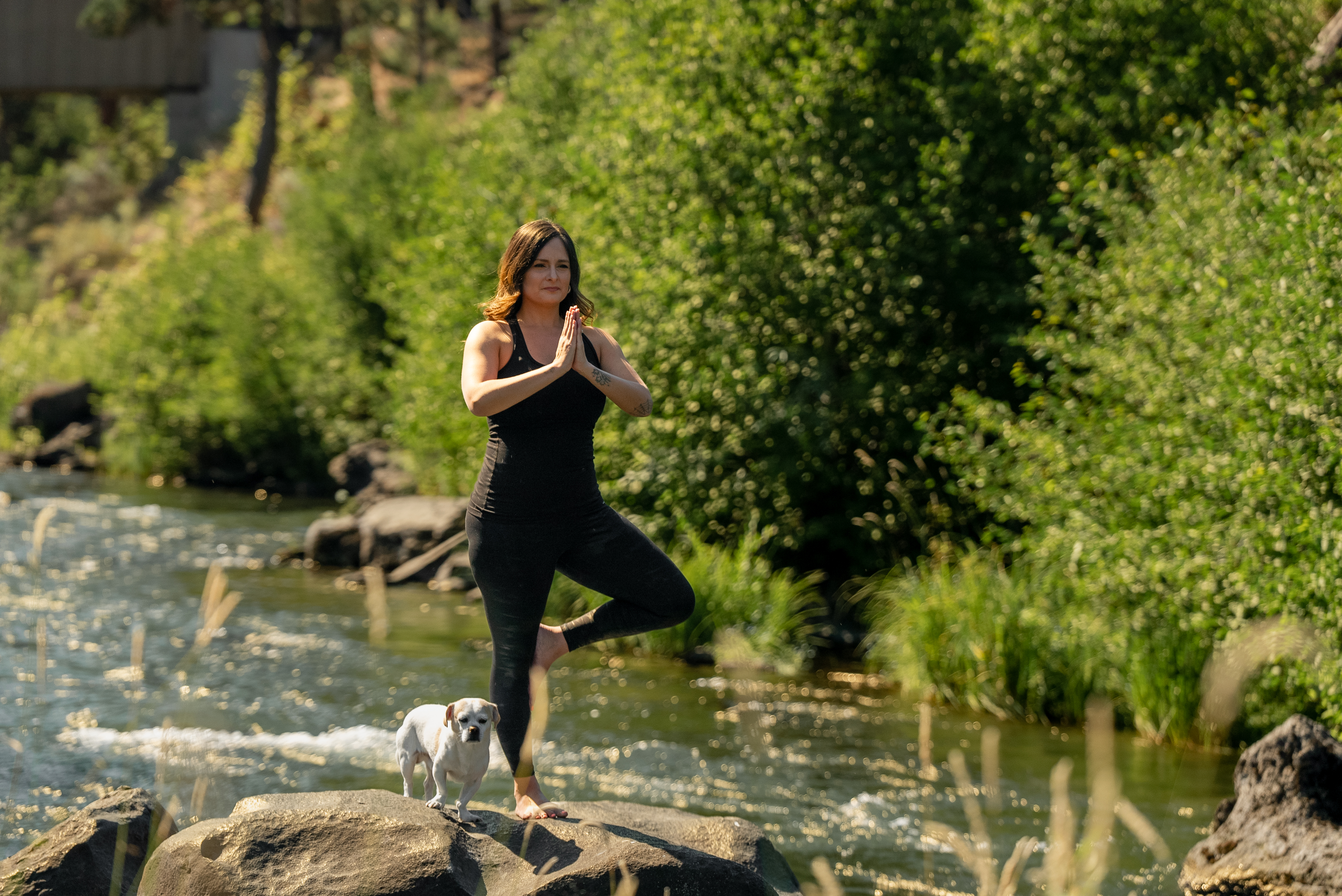 A woman is doing yoga on a rock by a river at Riverhouse Lodge in Bend Oregon; a dog stands beside her.