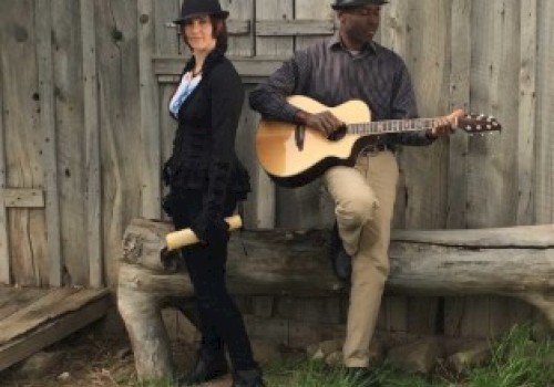 Two people wearing hats, with one holding a rolled paper and the other playing an acoustic guitar, are near a rustic wooden structure.