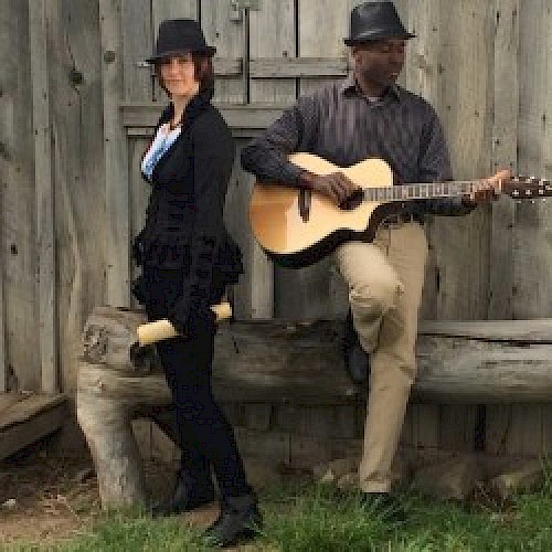 Two people wearing hats, with one holding a rolled paper and the other playing an acoustic guitar, are near a rustic wooden structure.