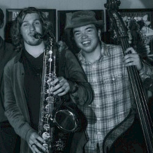 Two individuals, one holding a saxophone and the other a double bass, smile and pose for a photo in a black-and-white or sepia-toned image.
