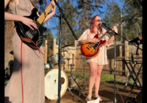 Two individuals are performing music outdoors with guitars, under a clear sky, surrounded by nature and musical equipment.