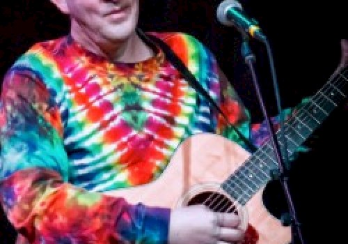 A person wearing a tie-dye shirt is playing an acoustic guitar on stage, standing in front of a microphone while performing.