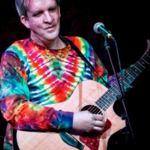 A person wearing a tie-dye shirt is playing an acoustic guitar on stage, standing in front of a microphone while performing.