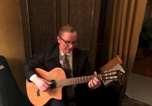 A man in a suit and tie is sitting and playing an acoustic guitar in a warmly lit room with screen dividers and a curtain in the background.