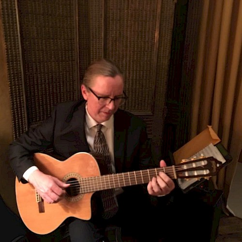 A man in a suit and tie is sitting and playing an acoustic guitar in a warmly lit room with screen dividers and a curtain in the background.