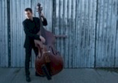 A person is playing a double bass in front of a corrugated metal wall, wearing a dark outfit and standing on a concrete surface.