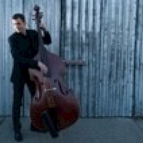 A person is playing a double bass in front of a corrugated metal wall, wearing a dark outfit and standing on a concrete surface.