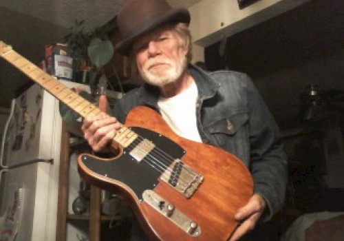 An older man with a beard is wearing a hat and jean jacket while holding an electric guitar inside a home with a refrigerator in the background.
