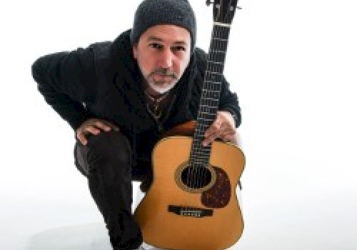 A person in a beanie and casual attire is crouching down while holding an acoustic guitar in front of them against a white background.