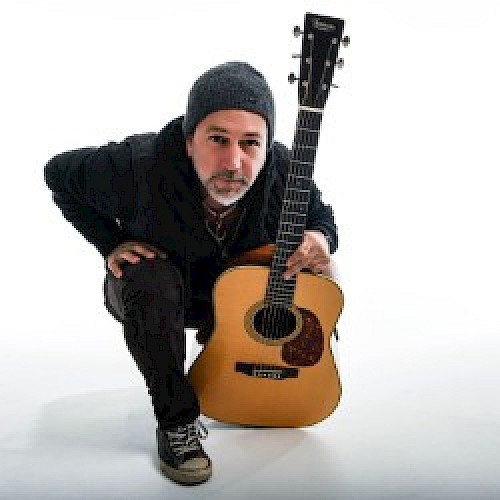 A person in a beanie and casual attire is crouching down while holding an acoustic guitar in front of them against a white background.