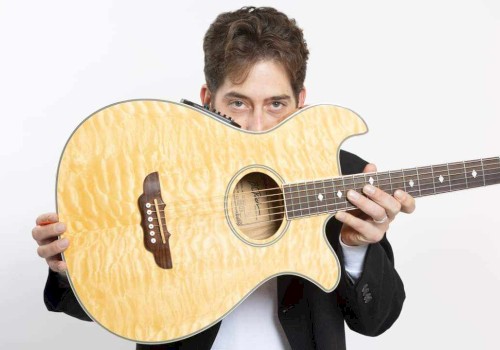 A person is holding a light-colored acoustic guitar, partially hiding their face behind it, with a white background.