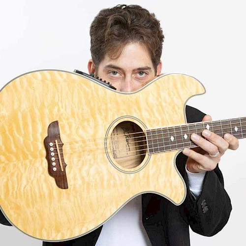 A person is holding a light-colored acoustic guitar, partially hiding their face behind it, with a white background.