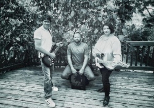 Three people are on a wooden deck, two holding guitars and one sitting down with greenery in the background.