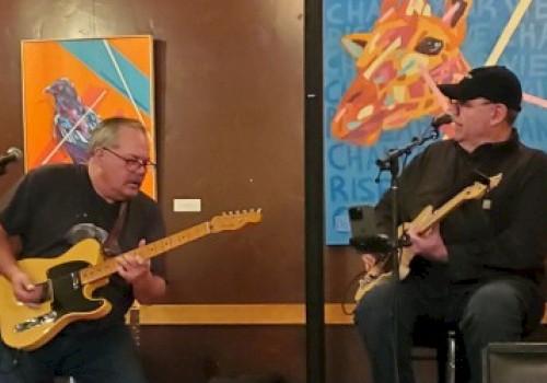 Two musicians are playing electric guitars in front of colorful paintings. One is standing while the other is seated.