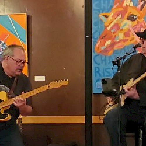 Two musicians are playing electric guitars in front of colorful paintings. One is standing while the other is seated.