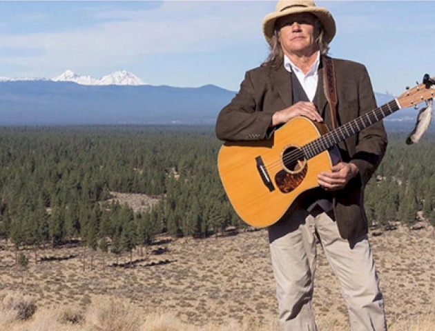 A person wearing a hat and holding an acoustic guitar stands in a vast open landscape with mountains and a forest in the background.