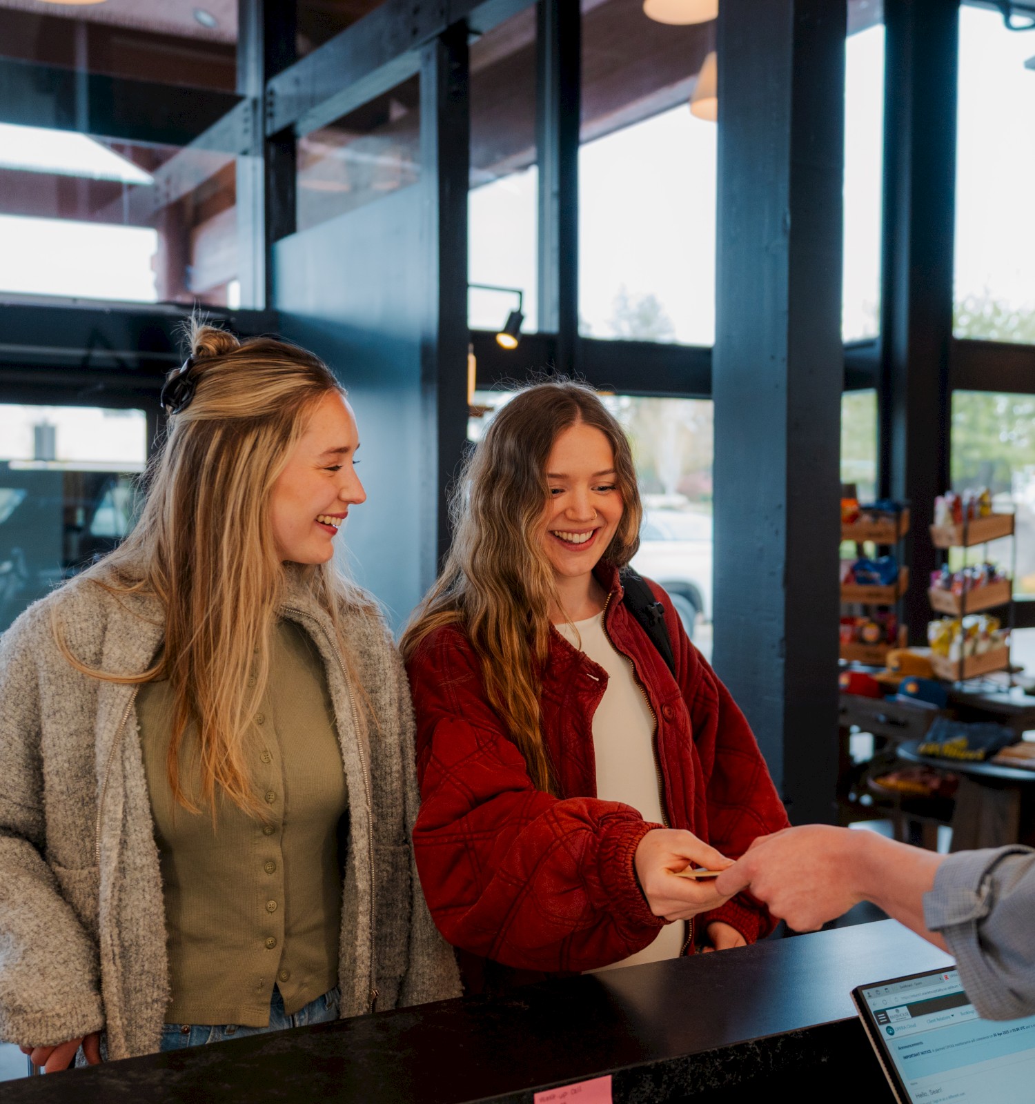 Two people checking in at Riverhouse Lodge reception desk in Bend, Oregon, smiling and handing over a key card. There's a lamp nearby and large windows in the background.