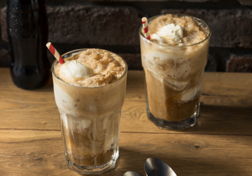 The image shows two root beer floats in glasses with striped straws, placed on a wooden table, with two spoons nearby and a bottle in the background.