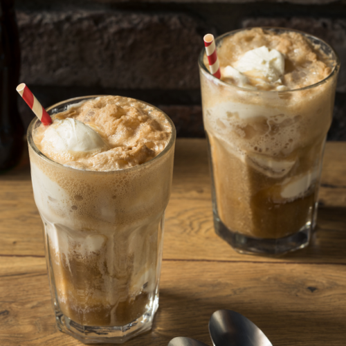 The image shows two root beer floats in glasses with striped straws, placed on a wooden table, with two spoons nearby and a bottle in the background.