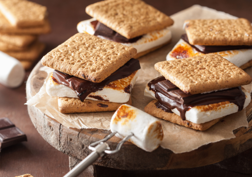 The image shows a wooden board with fresh s'mores made of graham crackers, toasted marshmallows, and chocolate, with extra ingredients around.