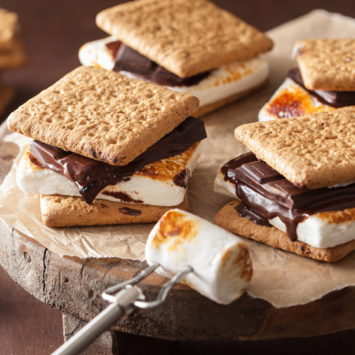 The image shows a wooden board with fresh s'mores made of graham crackers, toasted marshmallows, and chocolate, with extra ingredients around.