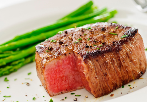 The image shows a perfectly cooked steak with a side of asparagus on a white plate, garnished with herbs and seasoning.