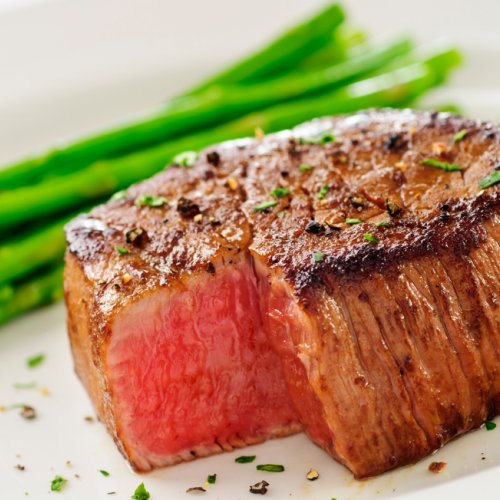 The image shows a perfectly cooked steak with a side of asparagus on a white plate, garnished with herbs and seasoning.