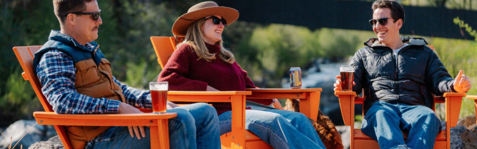 Three friends relax outdoors on orange chairs, enjoying drinks near a river with trees and a bridge in the background.