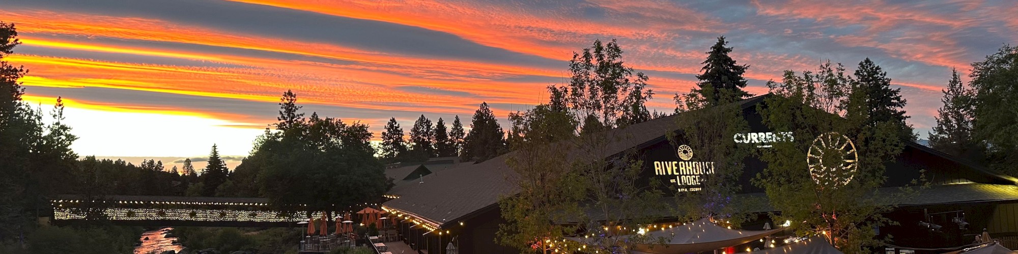 A scenic outdoor dining area by a river at sunset with string lights and colorful skies, surrounded by trees and mountains.