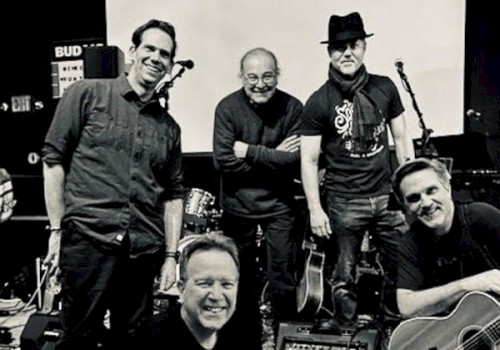 A black-and-white photo of five men posing with musical instruments in a stage setting, looking happy and relaxed.