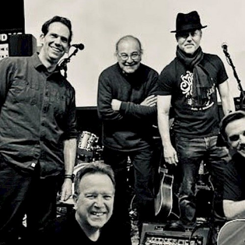 A black-and-white photo of five men posing with musical instruments in a stage setting, looking happy and relaxed.