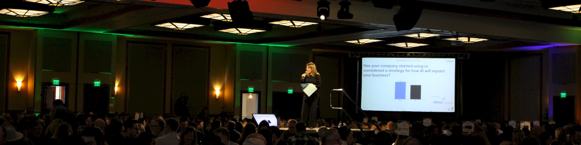 A person is speaking on a stage in a large conference room filled with attendees, with a presentation slide displayed behind them.