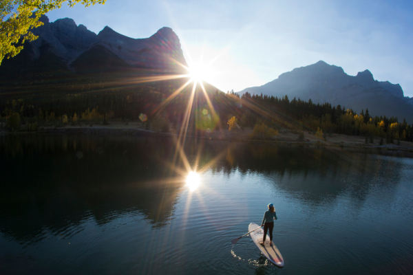 A person paddleboards on a tranquil lake surrounded by mountains, with the sun shining brightly and reflecting on the water's surface.