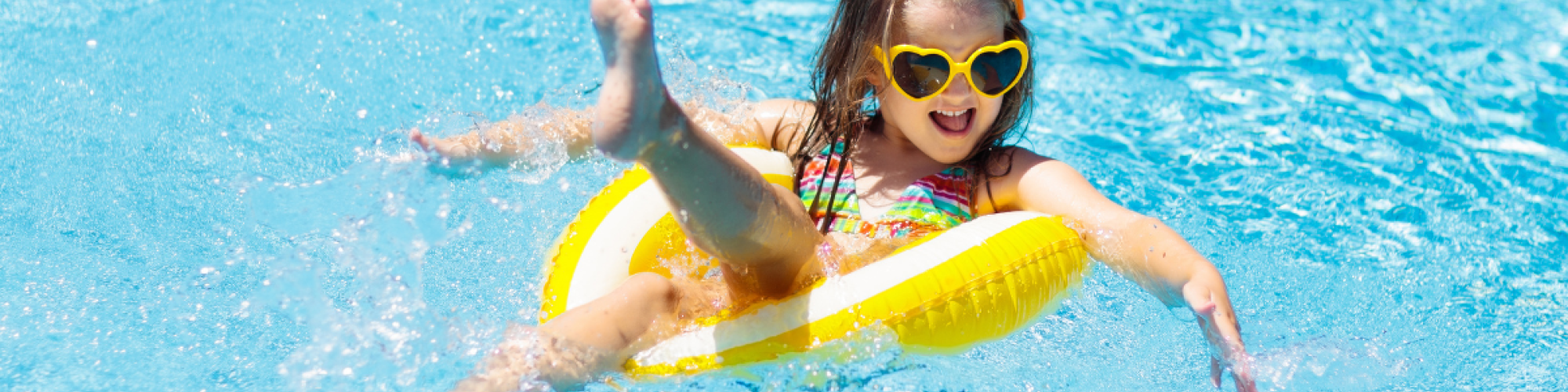 A child wearing heart-shaped sunglasses is happily playing in the outdoor pool at Riverhouse Lodge in Bend, Oregon, sitting in a yellow inflatable ring, splashing water around.