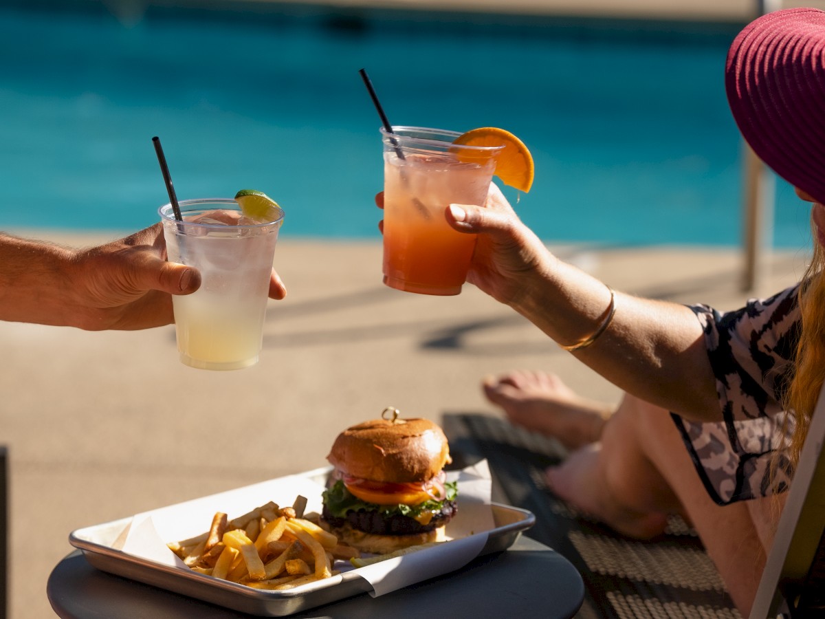 Two people clink drinks over a tray with a burger and fries, sitting by the outdoor pool at Riverhouse Lodge in Bend, Oregon, relaxing.