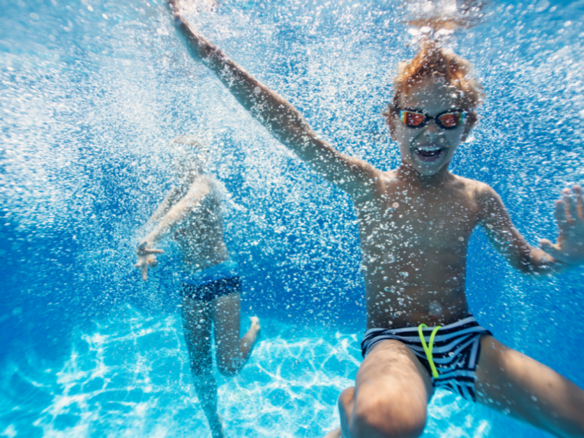 Two children are swimming underwater in Riverhouse Lodge outdoor pool in Bend, Oregon, one wearing goggles and smiling while the other swims in the background.