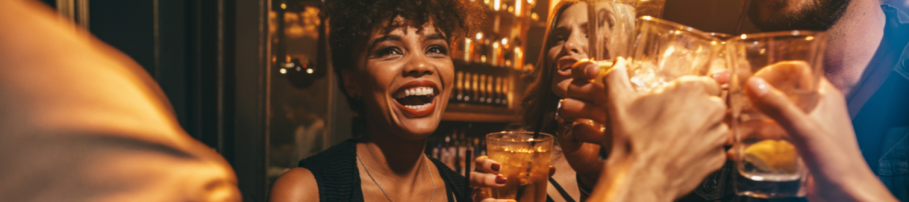 A group of friends is seen raising their glasses and celebrating in what appears to be a lively bar or club, filled with warm lighting and cheerful vibes.