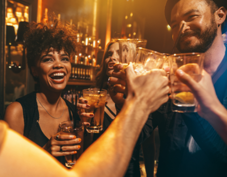 A group of friends is seen raising their glasses and celebrating in what appears to be a lively bar or club, filled with warm lighting and cheerful vibes.