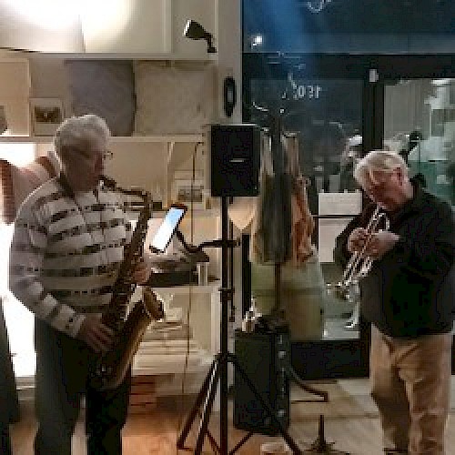 Two musicians are playing instruments indoors, one on a saxophone and the other on a trumpet, with a microphone and music stand in between them.