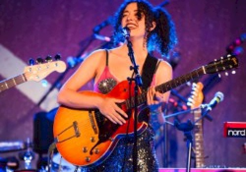 A musician is performing on stage, playing an electric guitar, with a microphone in front and musical equipment around her, under a blue 