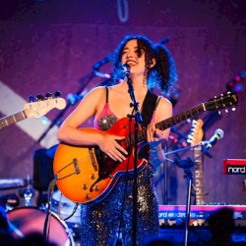 A musician is performing on stage, playing an electric guitar, with a microphone in front and musical equipment around her, under a blue 